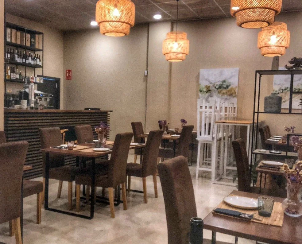 A cozy restaurant interior with wooden tables, brown upholstered chairs, and decorative hanging lamps. The tables are set with plates, glasses, and small flower arrangements. The background features a bar counter, shelving with bottles, and wall art depicting a landscape scene.