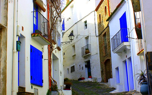 A narrow street in a Mediterranean town with white buildings, some with blue shutters and doors, and small balconies. The street is paved with cobblestones and has a slight incline.