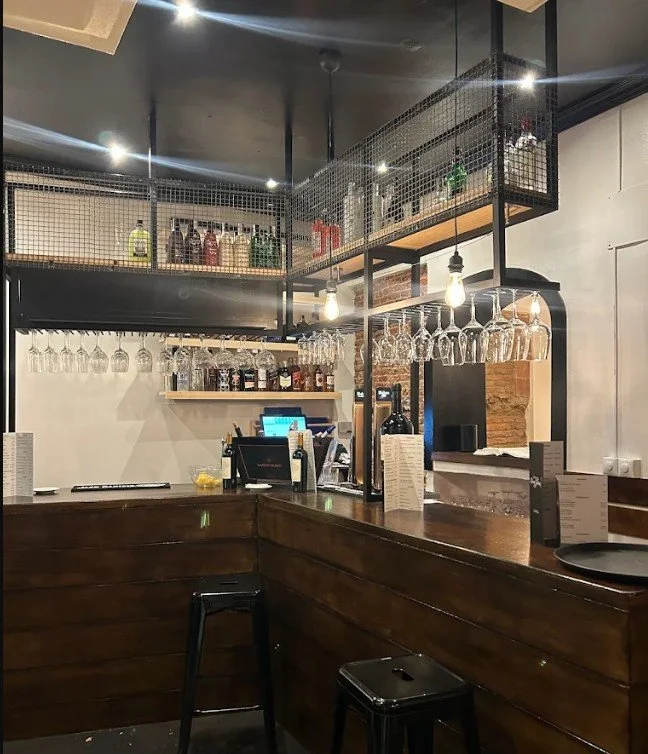 Interior of a modern bar with hanging wine glasses, bottles, and illuminated pendant lights over a wooden counter.