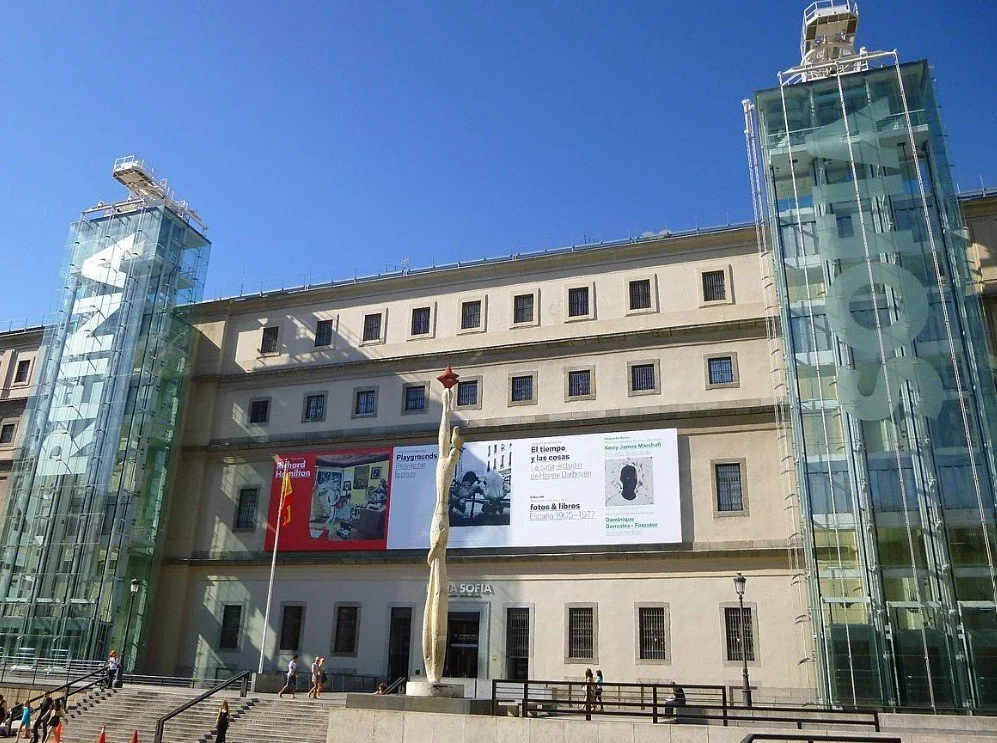 Exterior of a modern building with glass elevators on each side and banners. In front, a sculpture and people walking.
