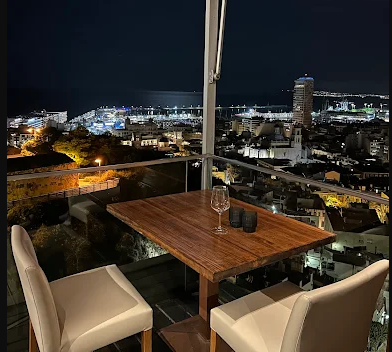 Nighttime city view from a balcony with a wooden table, a glass, and two cream-colored chairs.