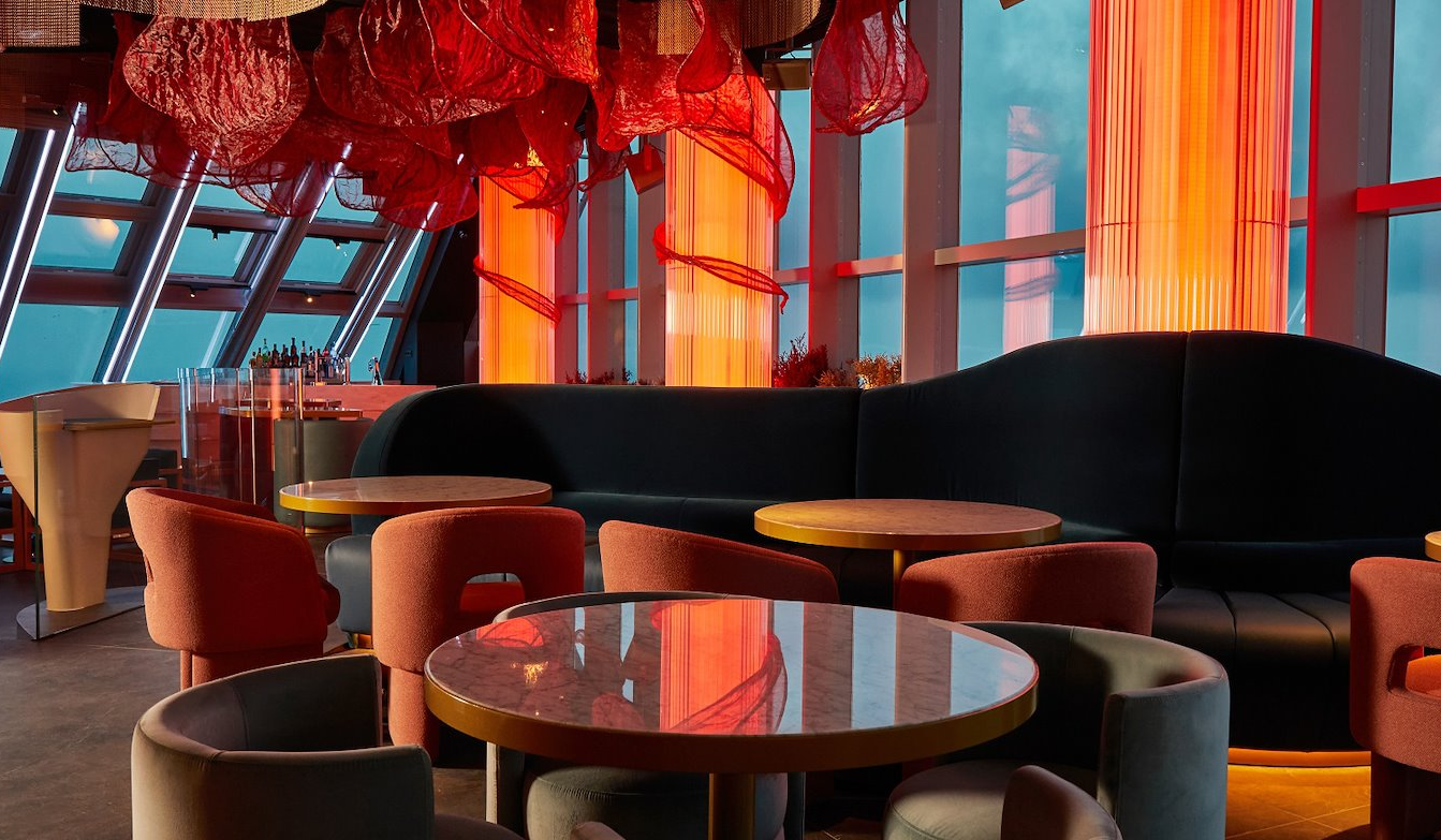 Interior of a modern lounge or restaurant with round tables and assorted chairs, large vertical orange light fixtures, a black curved sofa, and decorative orange fabric hanging from the ceiling, with floor-to-ceiling windows showing a blue sky.