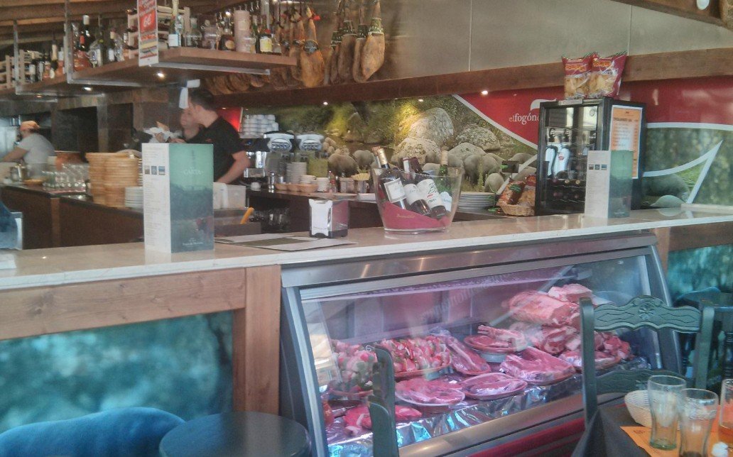 Interior of a restaurant or butcher shop with a meat display case filled with various cuts of raw meat. A counter with bar stools, bottles, and glasses is visible, along with shelves of bottles and containers behind the counter. Customers and staff are present in the background.