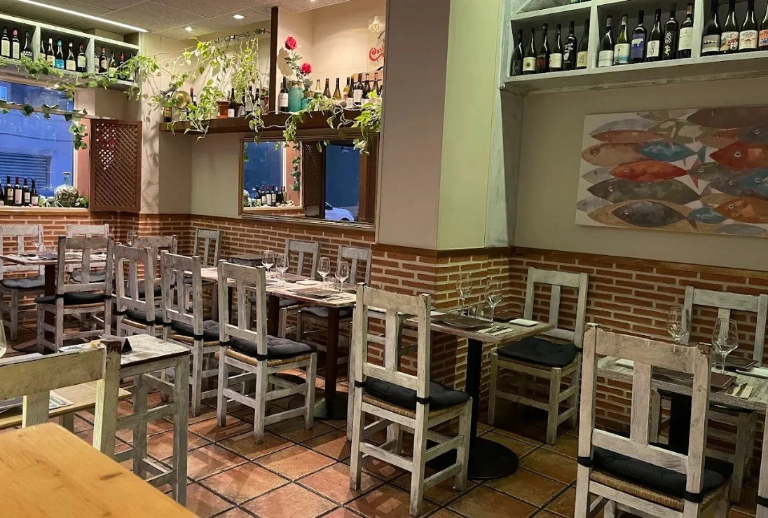 Interior of a cozy restaurant with rustic wooden chairs, wine bottles on shelves, and a fish-themed painting on the wall.