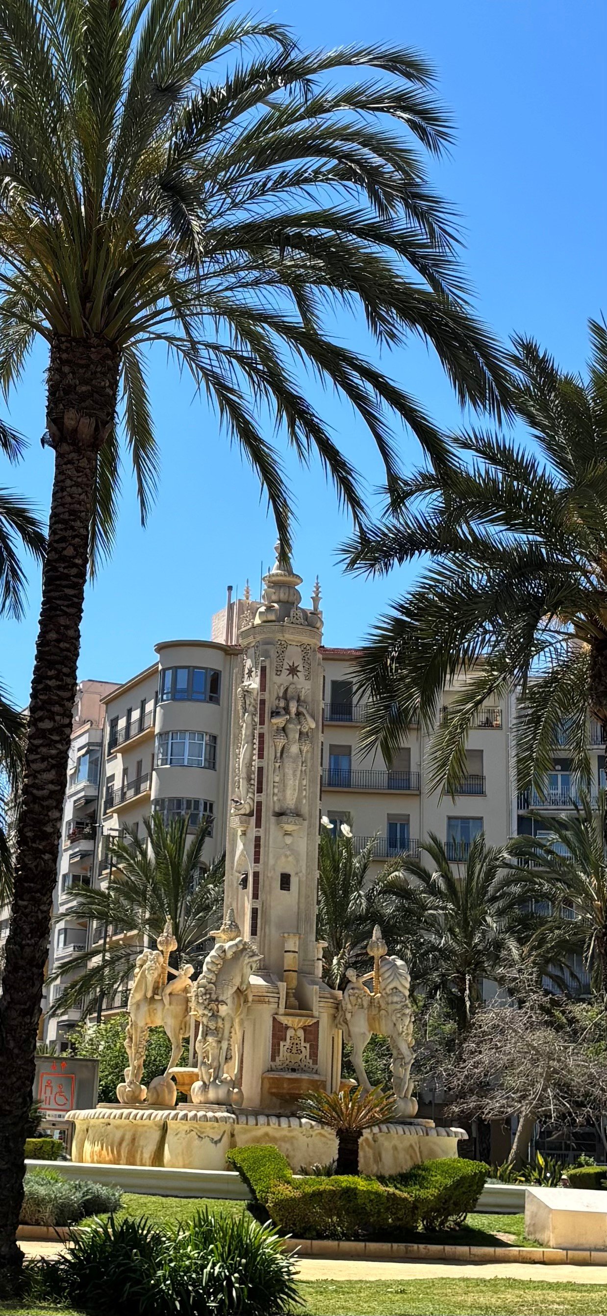 A decorative fountain with sculptures and a tall ornate building with balconies, framed by tall palm trees under a bright blue sky.
