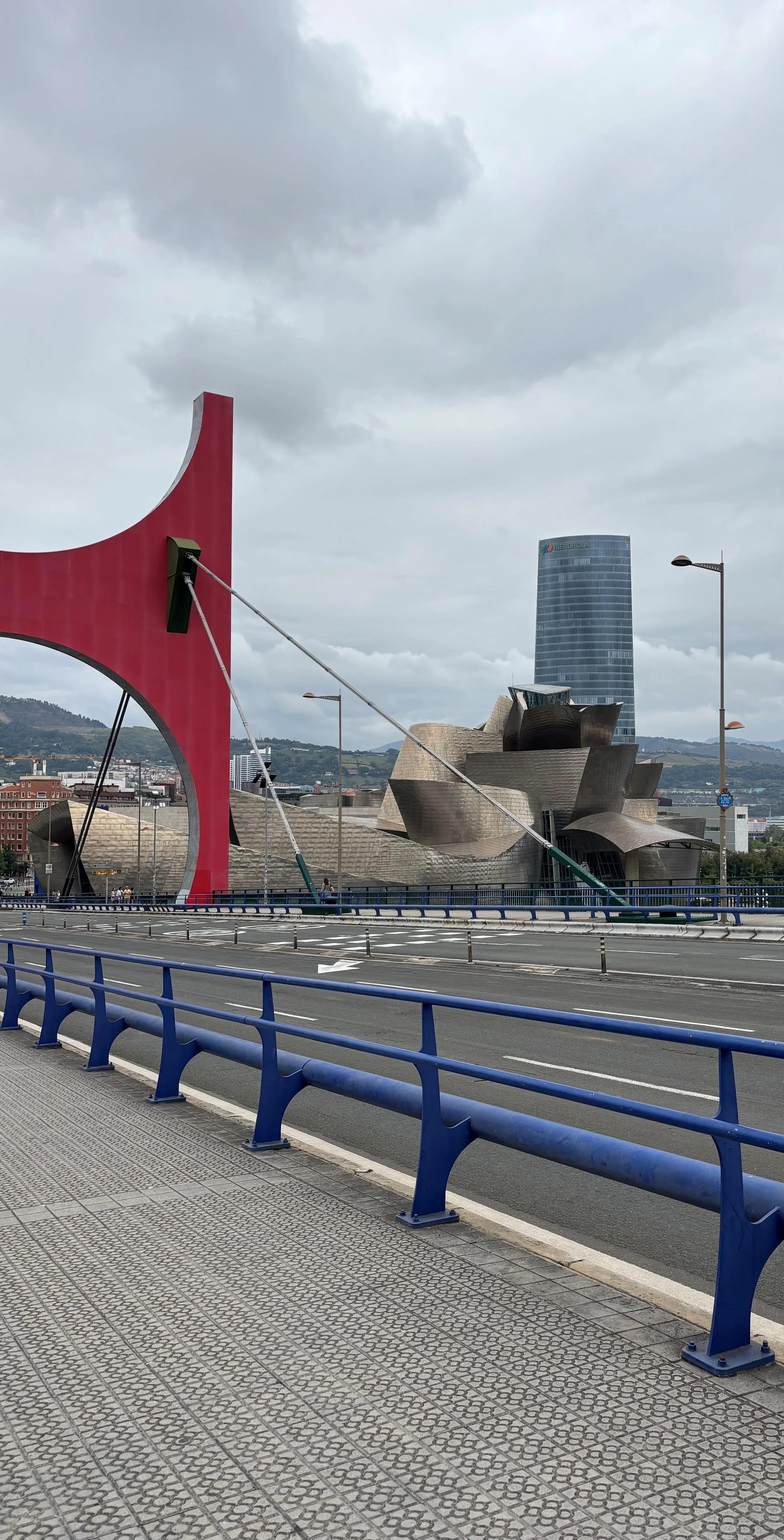 View of Bilbao, Spain with the Guggenheim Museum in the foreground, a modern curved building made of titanium, and a tall glass skyscraper in the background on a cloudy day.