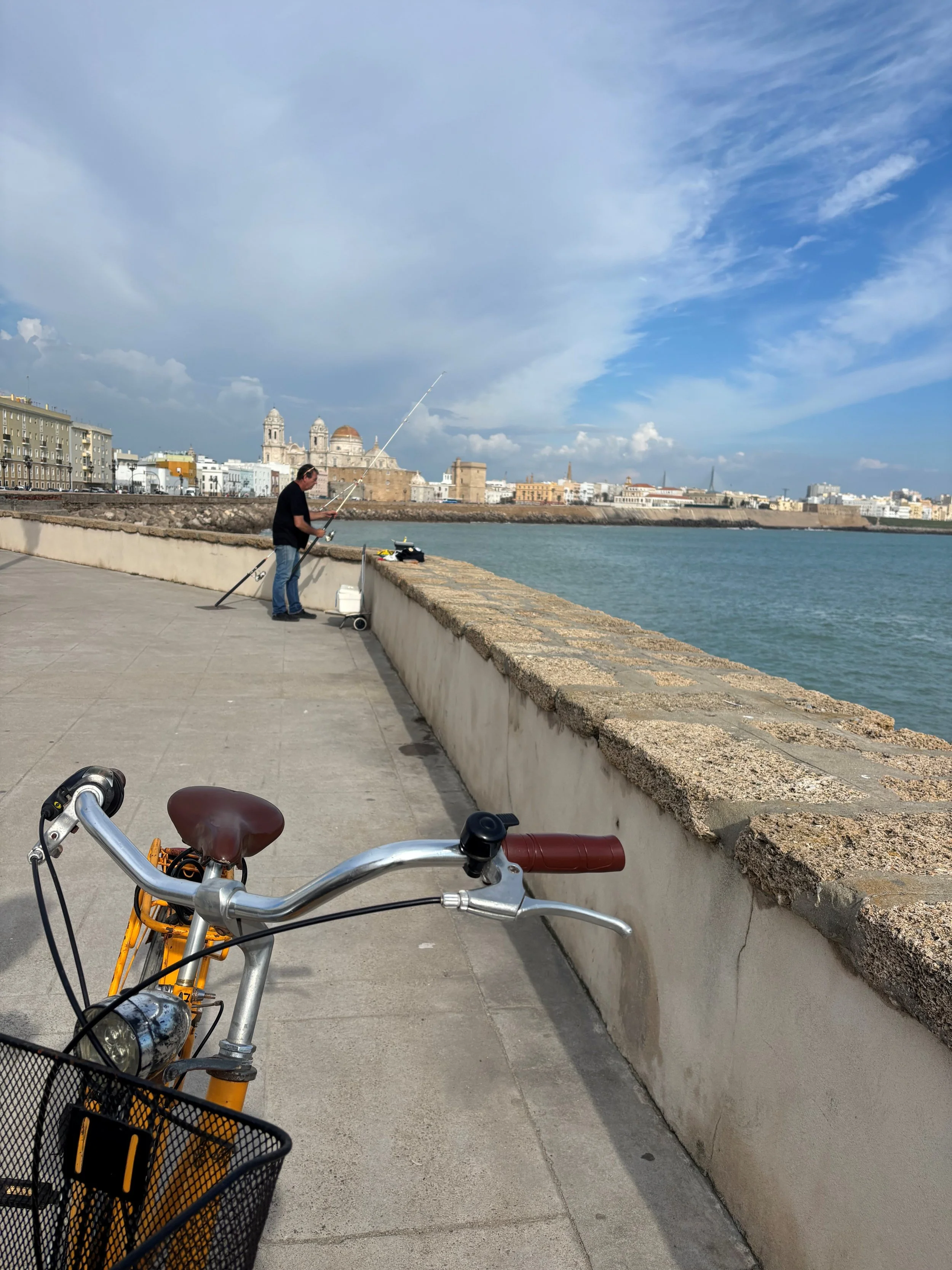 A man fishing along a historic waterfront with city buildings and a large cathedral in the background. Bicycles are in the foreground.