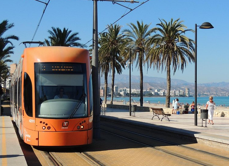 An orange tram on tracks alongside a beach promenade with people walking and sitting on benches, palm trees, and the ocean in the background.