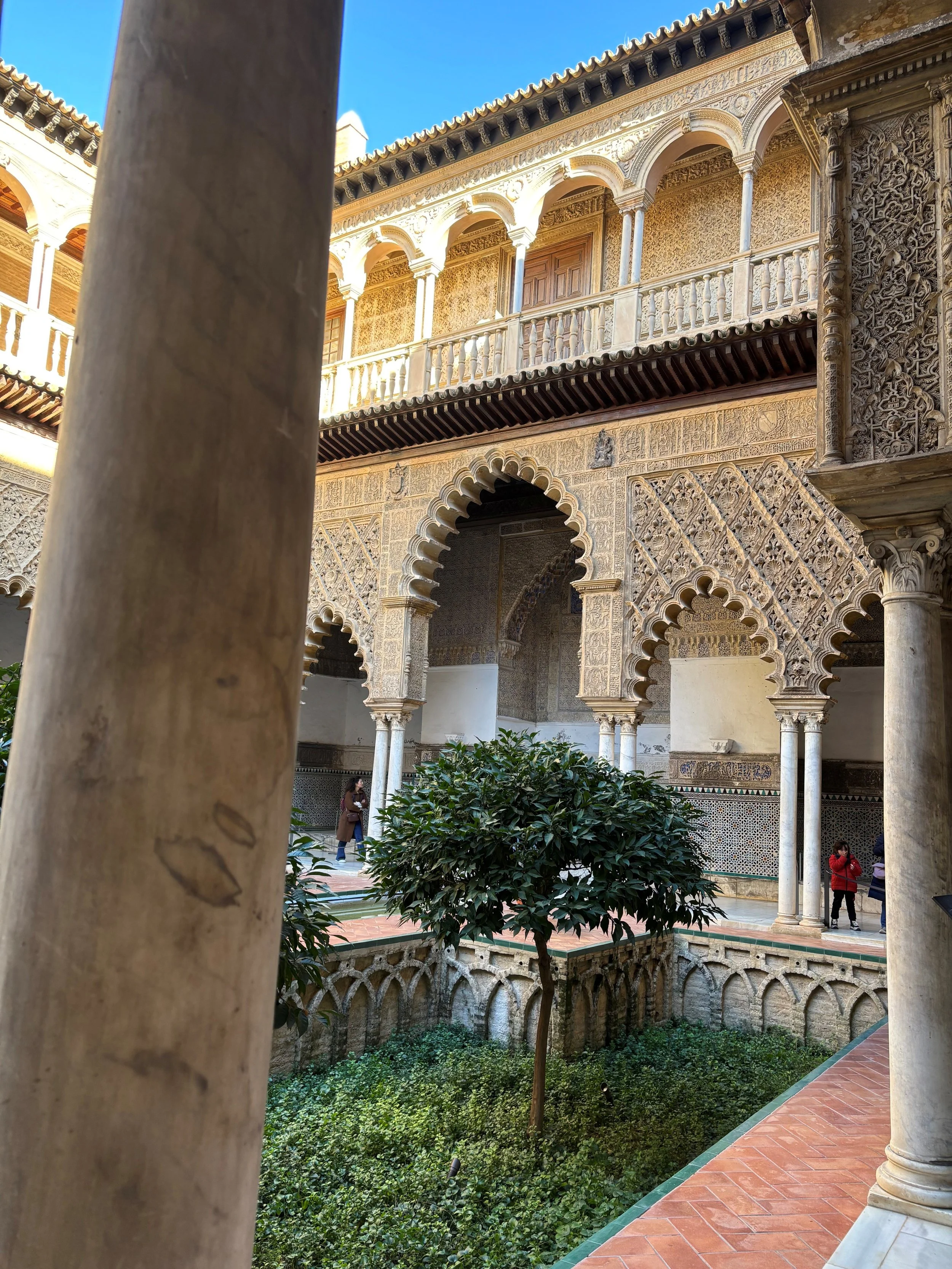 Courtyard with traditional Islamic architecture, intricate carvings on walls, arched doorways, columns, green plants, and visitors taking photos.