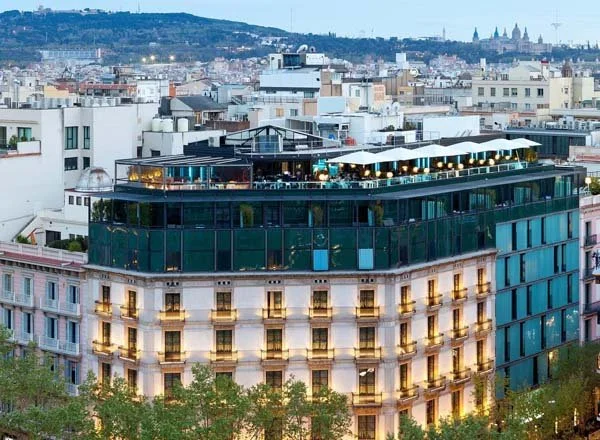 A multi-story building in an urban setting with a rooftop terrace featuring seating and lighting, surrounded by other buildings with distant cityscape and hills.