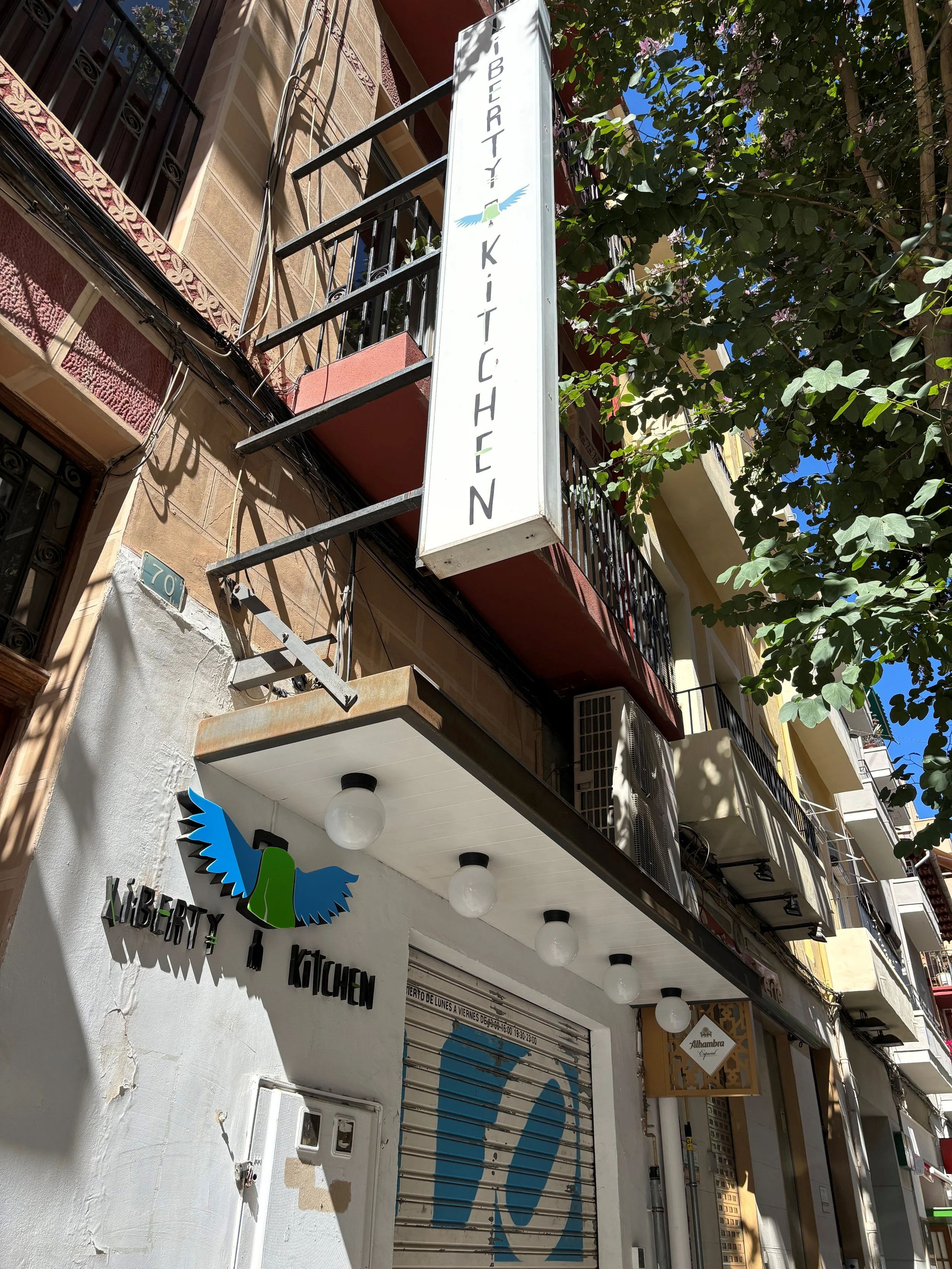 Street view showing a white building with balconies, a sign that reads 'Liberty Kitchen', a logo with a blue eagle, and storefronts with metal shutters, decorative lights, and trees lining the street under a clear blue sky.