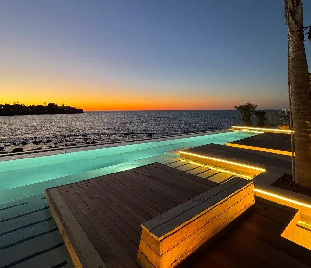A poolside area overlooking the ocean during sunset with built-in lighting and wooden lounge chairs.