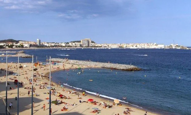 Beach with people sunbathing and swimming, cityscape in the background, blue sky with a few clouds.