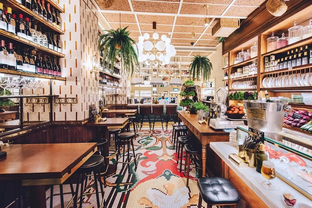 Interior of a cozy cafe or restaurant with wooden tables, bar stools, shelves filled with bottles and jars, hanging plants, decorative mosaic tile flooring, and soft lighting.