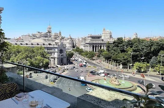 Cityscape view showing a busy street with cars, historical buildings, green trees, and a roundabout with a fountain, taken from a high vantage point on a balcony.