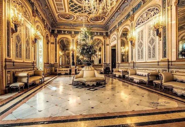 Luxurious hotel lobby with ornate gold and white decor, chandeliers, marble floors, and vintage-style seating arrangements.