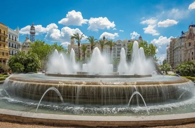 A large circular fountain with multiple water jets in a city square, surrounded by historic buildings, palm trees, and a partly cloudy sky.