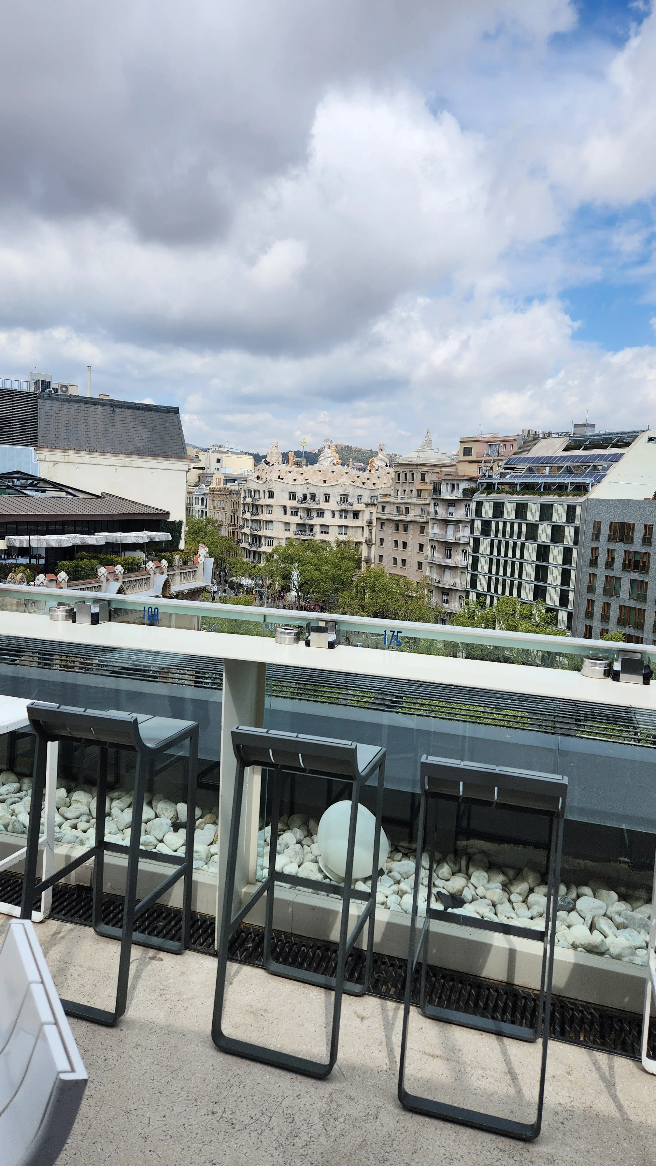 Cityscape view from a terrace with chairs, railing, and rocks, overlooking buildings with cloudy sky.