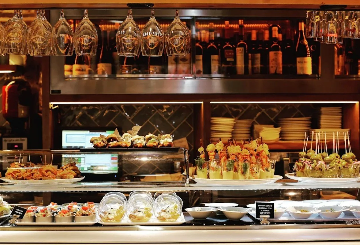 Display of assorted appetizers and desserts in a restaurant buffet, with wine glasses hanging overhead and wine bottles on shelves in the background.