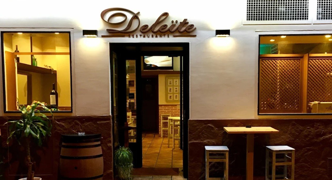 Exterior view of a restaurant called 'Deliite,' showing the entrance with a sign, two windows, and some outdoor furniture including a small barrel, a plant, and a table with two stools.