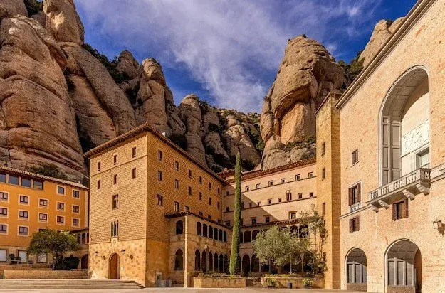 Historic monastery complex built into rocky cliffs with stone buildings, arched windows, and a courtyard, under a partly cloudy sky.