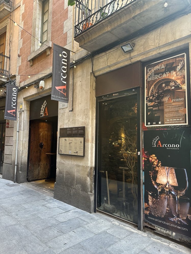 Entrance of a restaurant called Arcano with a black glass door, outdoor signage, and a menu display, located on a city street with stone pavement.