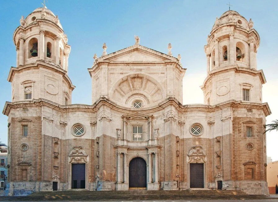 Front view of a historic baroque-style church with two bell towers, intricate architectural details, and a central arched entrance, set against a clear blue sky.