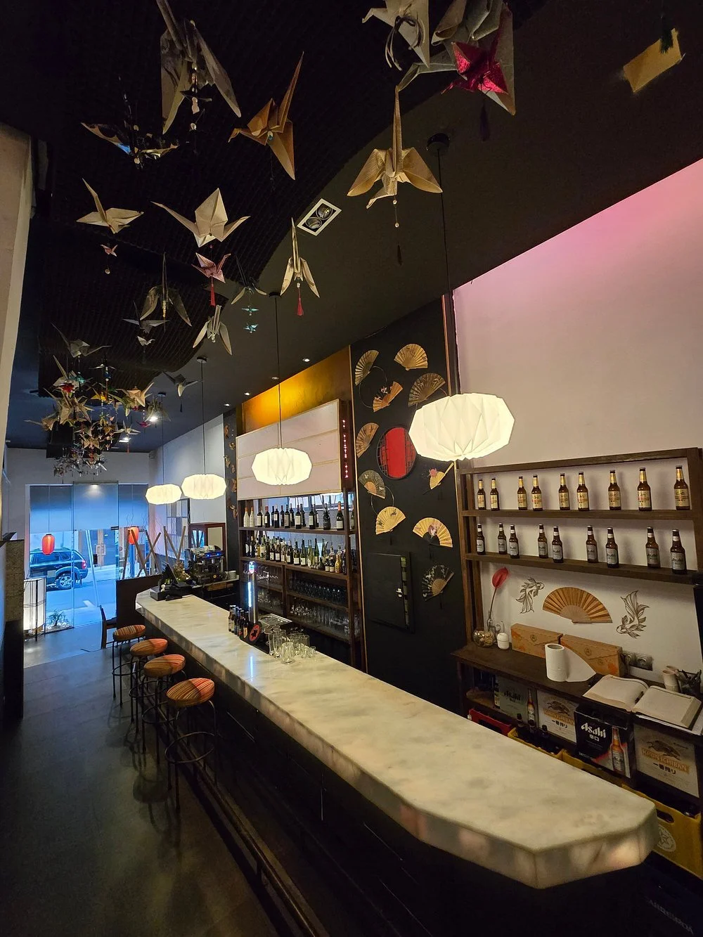 Interior of a bar with origami cranes hanging from the ceiling, illuminated white pendant lights, shelves with bottles of alcohol, and a marble-topped counter with bar stools.