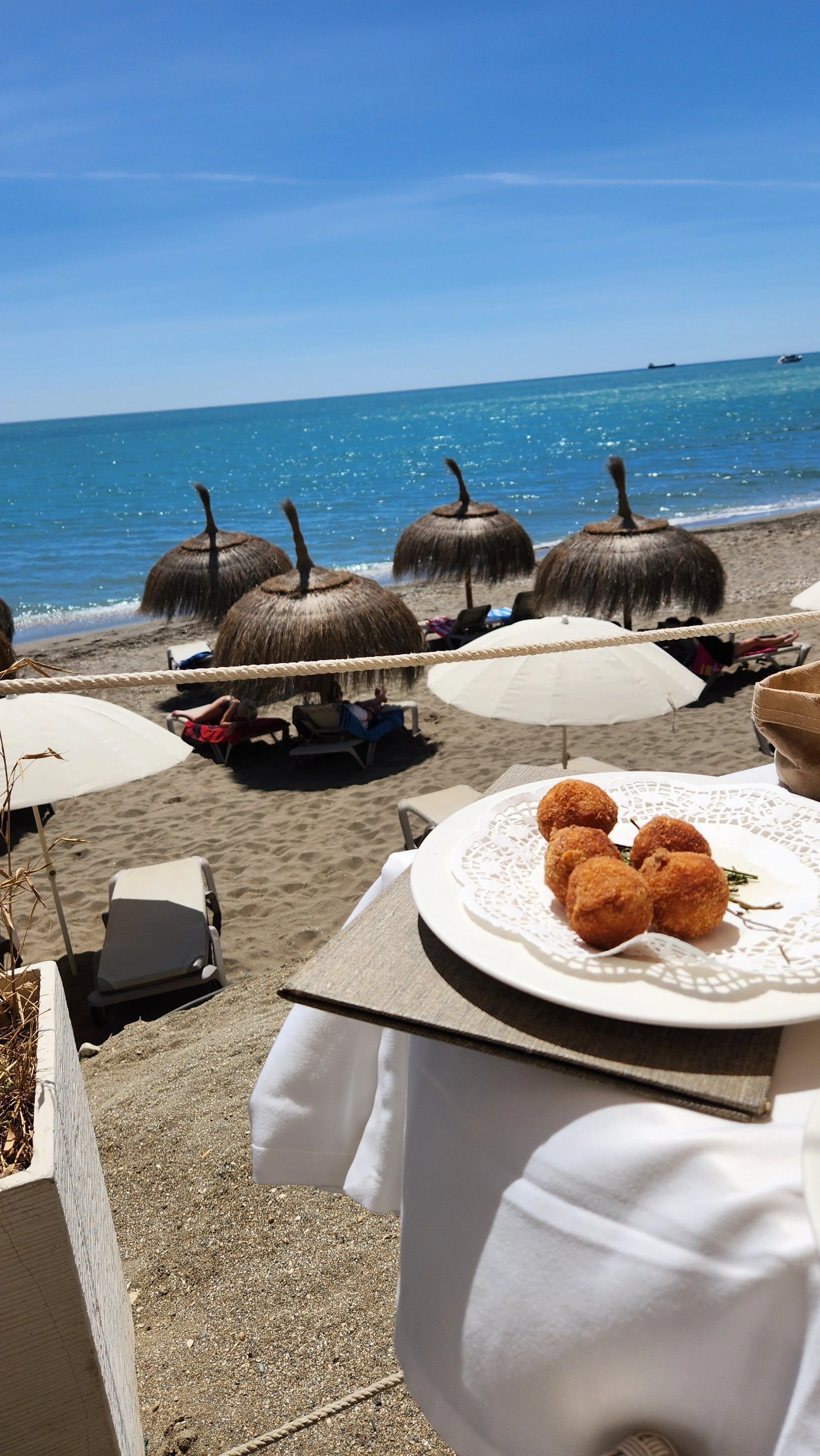 Plate of fried food on a table at the beach, with straw umbrellas, lounge chairs, and people relaxing near the water, and ships on the ocean in the background.