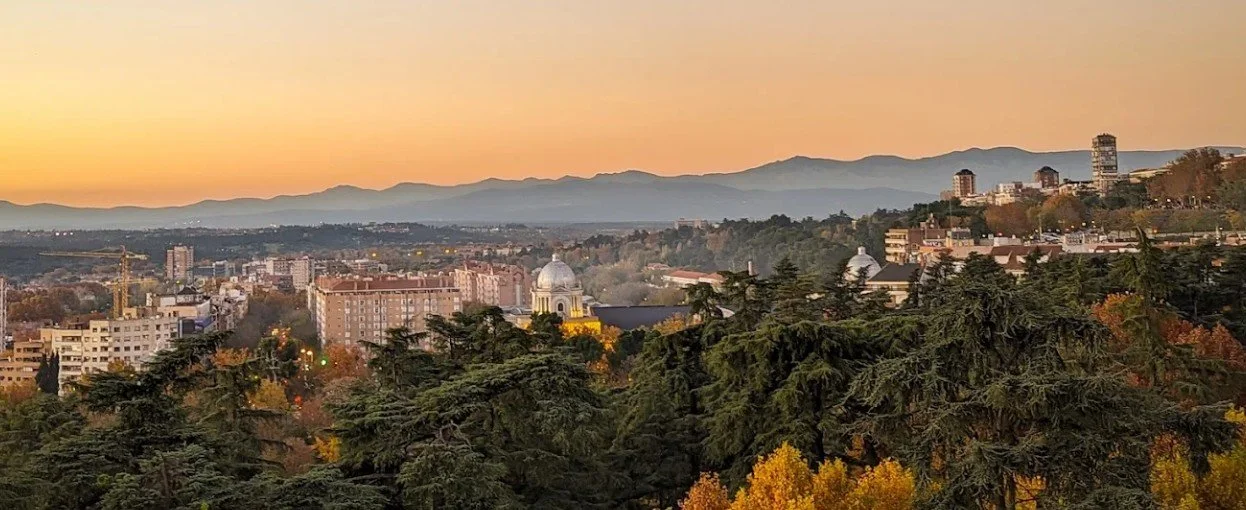 Cityscape at sunset with mountains in the background, buildings, and a foreground of trees with autumn foliage.