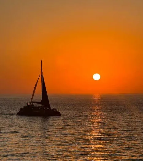 A sailboat on the ocean during sunset with an orange sky and the sun near the horizon.