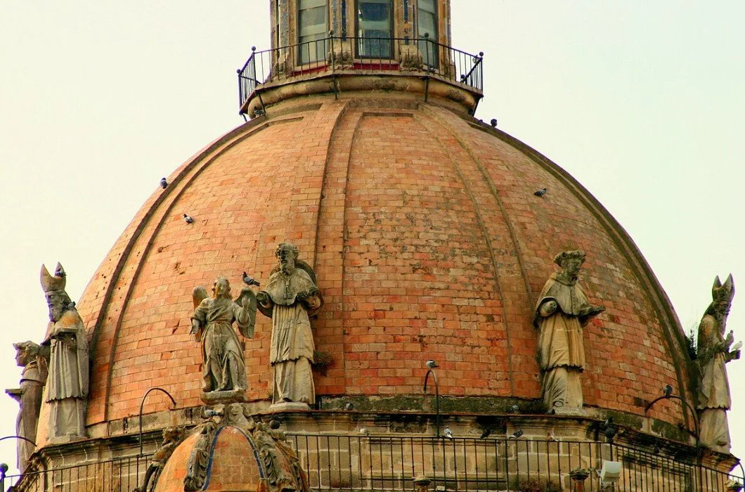 Close-up of an old brick domed building with sculptures of people and animals on its surface, with a railing at the top.