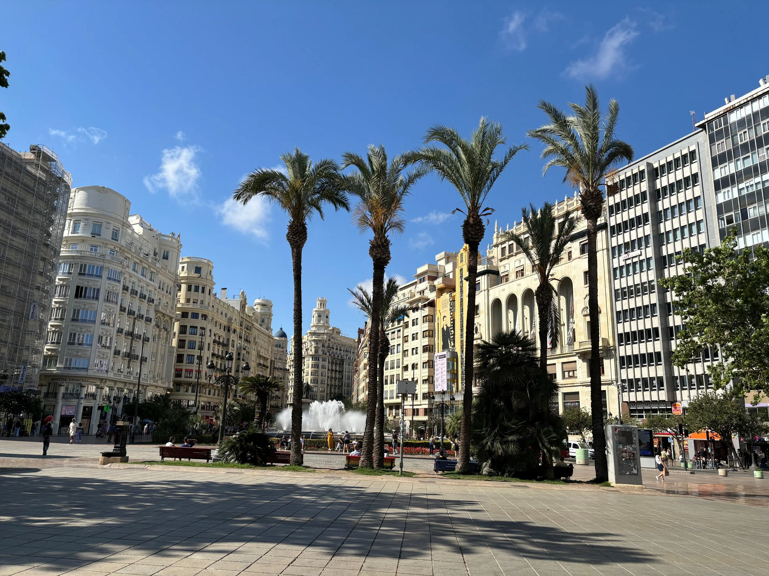 City square with palm trees, historic buildings, a fountain, and people walking.