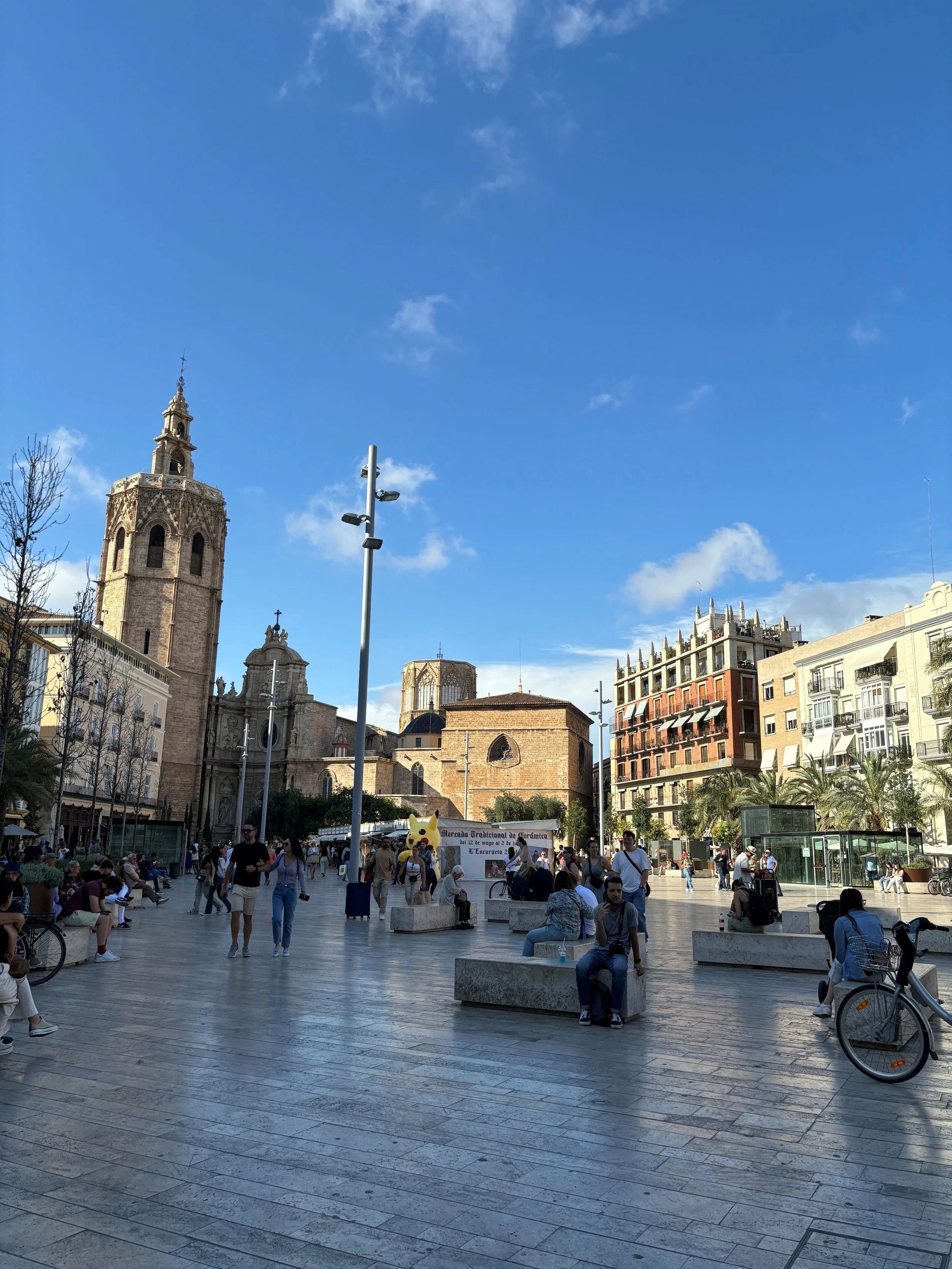 A busy city square with people sitting on benches, walking, and gathering. There are historic buildings, a church with a tall tower, and modern structures with palm trees lining the area. The sky is clear with a few clouds.