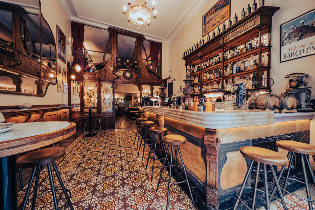 Elegant bar with vintage decor, wooden furniture, bar stools, shelves with bottles, ornate tiles on floor, and warm lighting.