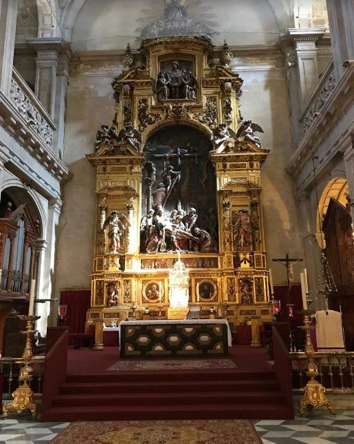 An ornate religious altar inside a church, featuring multiple sculptures depicting a biblical scene, gilded decorations, and a central cross, with steps leading up to it and a patterned rug in front.