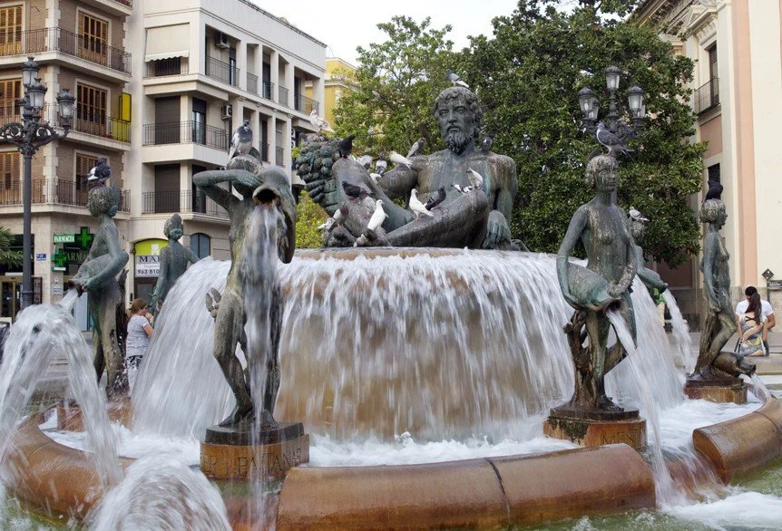 A fountain with a large statue of a bearded man and several smaller child-like figures, with water flowing from the fountain, in an urban setting surrounded by buildings and people.