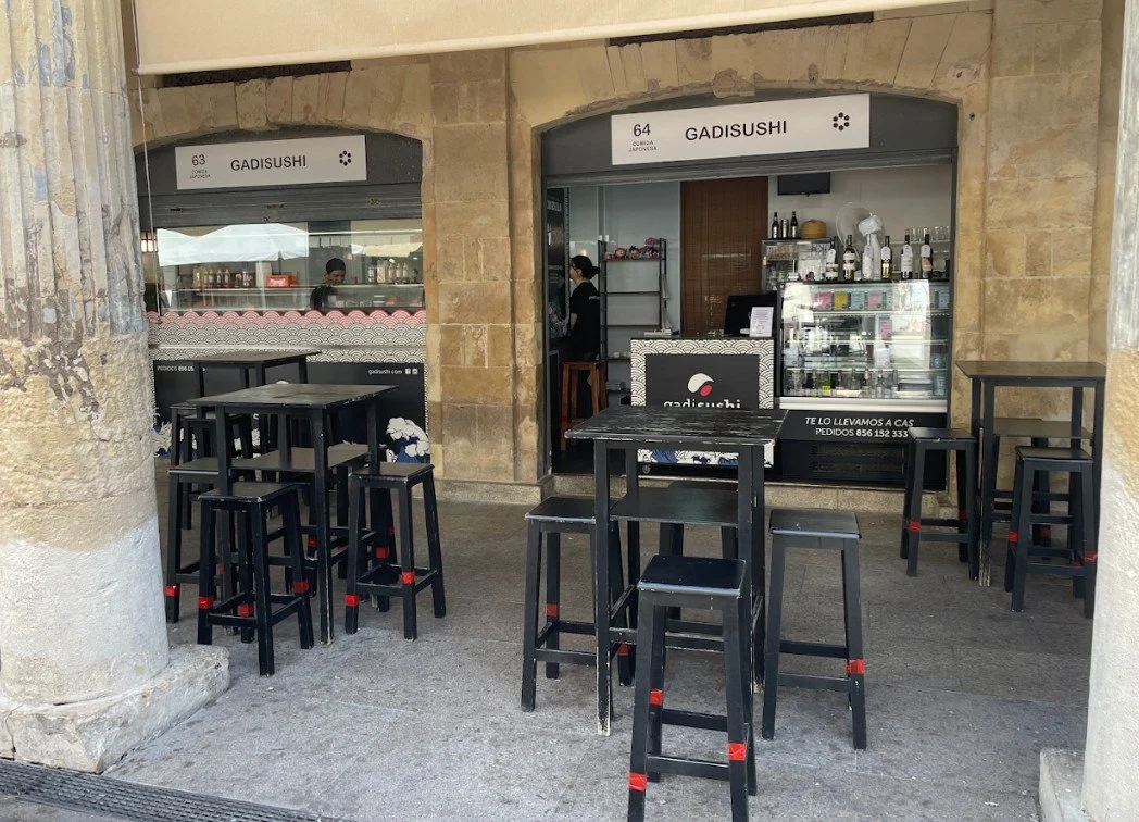 Outdoor seating area with black tables and high stools outside a sushi restaurant called GADISUSHI, located inside a building with stone walls.