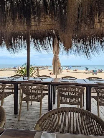 View of a beachside restaurant with wooden tables and chairs, thatched roof overhead, and the beach with umbrellas and people in the background.