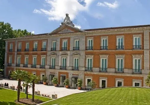 A large historic building with classical architecture, a triangular pediment, and a statue on top, surrounded by landscaped grass and palm trees, under a blue sky with some clouds.