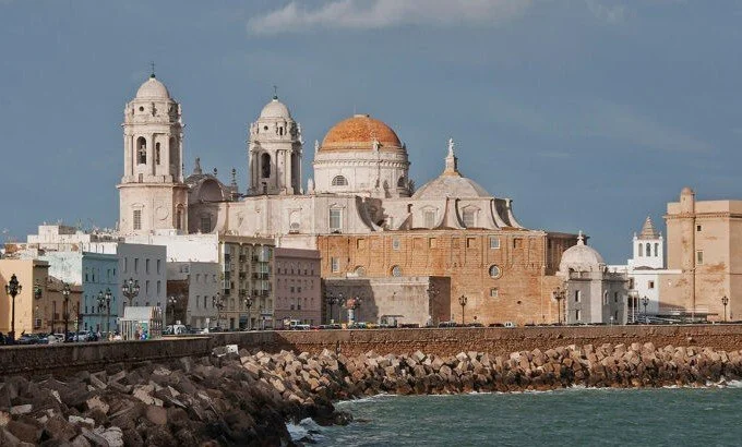 Historic coastal cityscape with a stone pier, featuring a large cathedral with domes and bell towers, and older multi-story buildings along the shoreline, under a partly cloudy sky.