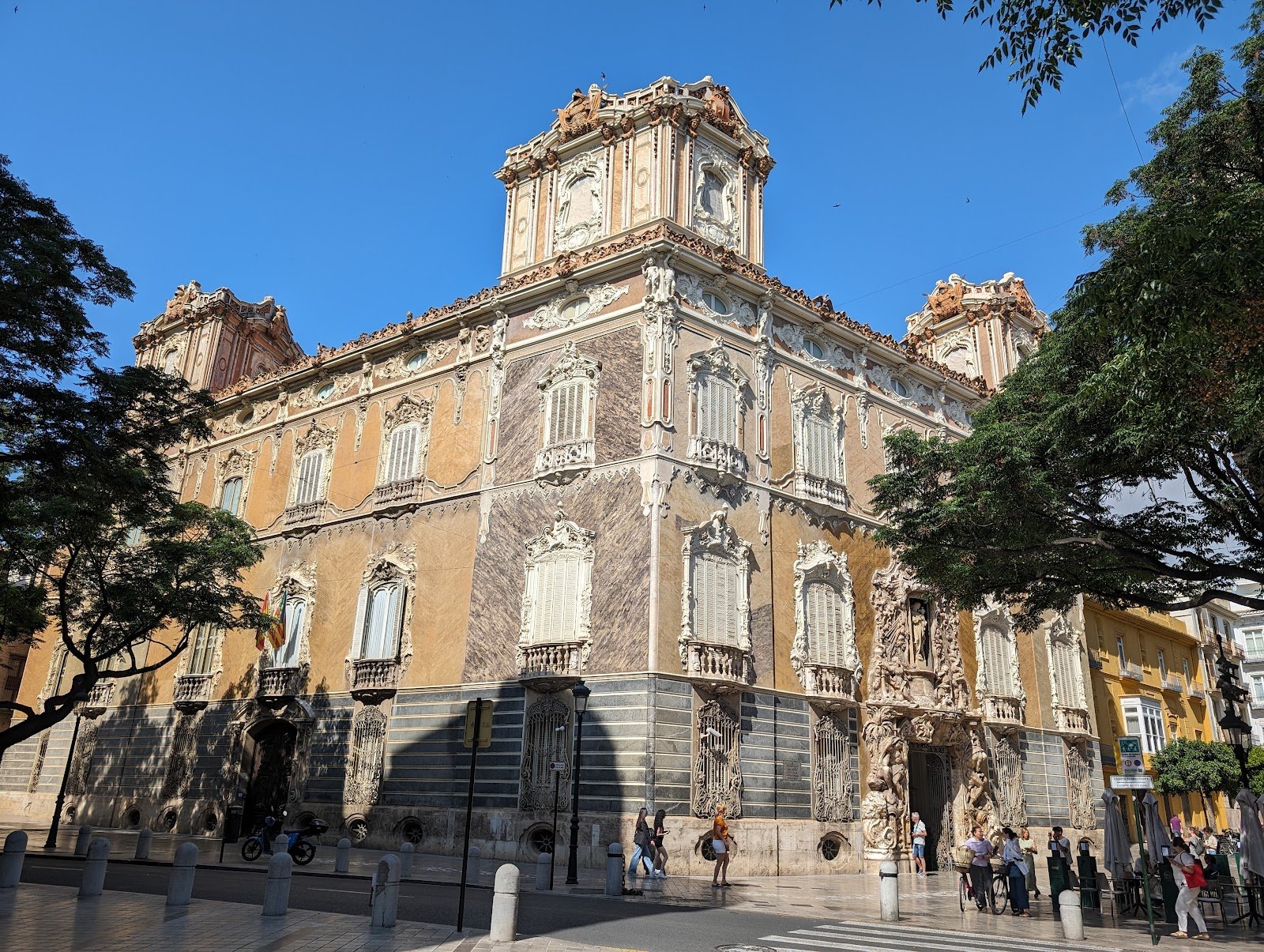 Historical building with ornate architectural details, including decorative window frames and sculptures, on a city street with pedestrians and trees.