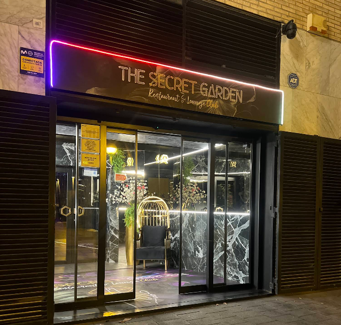Night view of the entrance to The Secret Garden, a restaurant and lounge club, with glass doors, a black marble wall inside, a black chair with a gold birdcage decor, and neon signs.