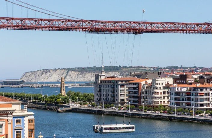 View of a city waterfront with a suspension bridge overhead, high-rise buildings along the water, and a cable car in the water.