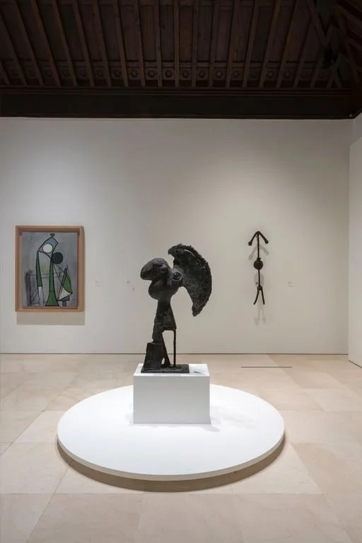 An art gallery features three sculptures and one painting. The central sculpture on a circular white platform depicts a stylized figure with a large, rounded head and abstracted body parts. To the left, a colorful abstract painting with geometric shapes hangs on the white wall. To the right of the sculpture, a slim, elongated hanging sculpture resembling a person is displayed. The gallery has a wooden ceiling with visible beams and a polished stone floor.