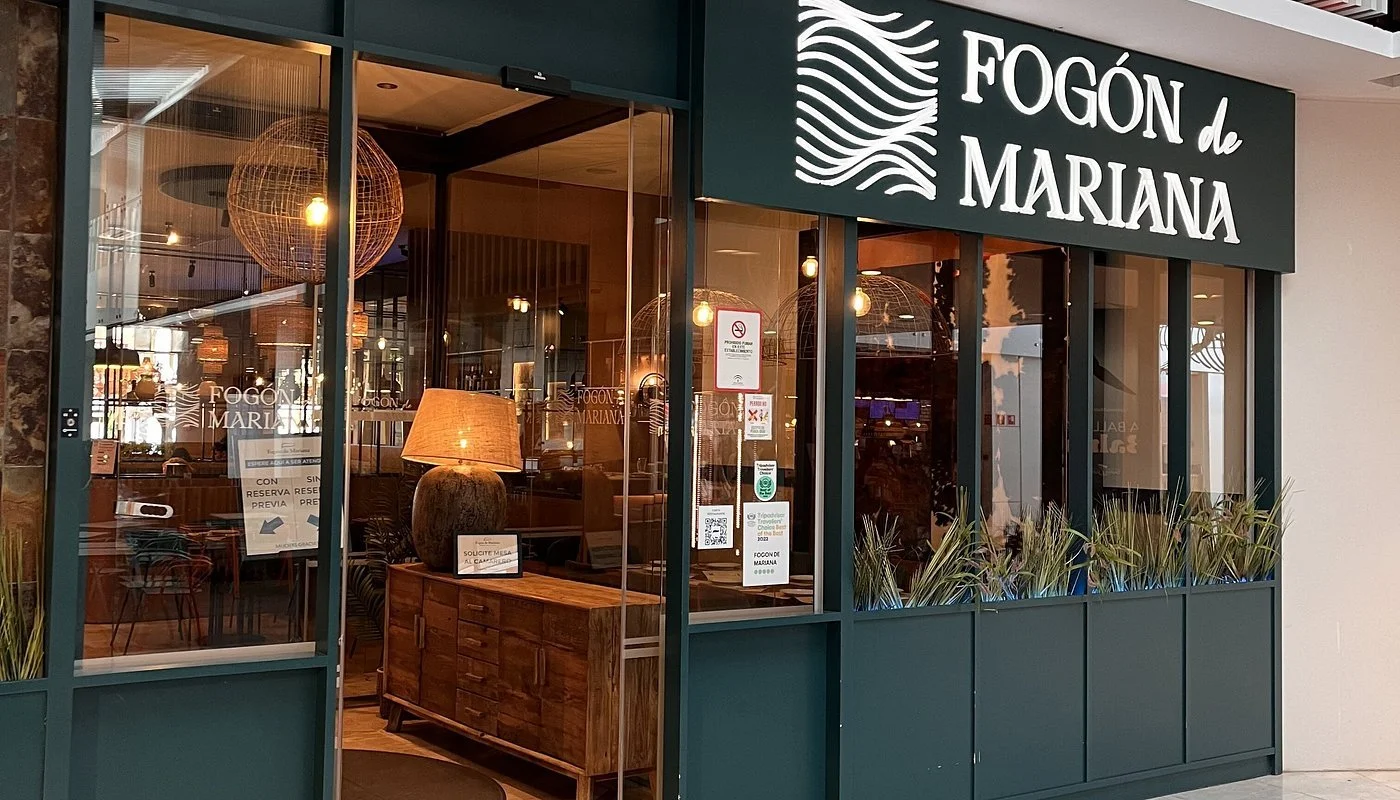 The storefront of Fogón de Mariana restaurant with large glass windows displaying the interior, which has warm lighting and wooden furniture, and the restaurant's name on a large sign above the entrance.