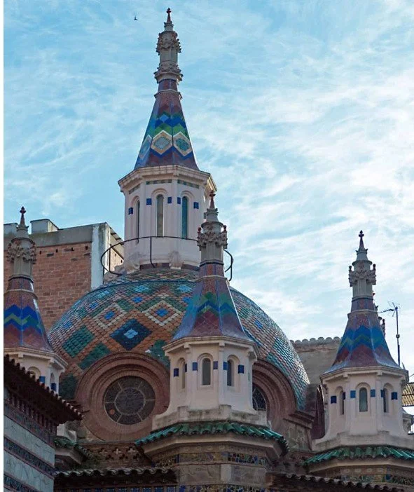View of a colorful, ornate church roof with four spires decorated with geometric patterns and a cloudy sky in the background.
