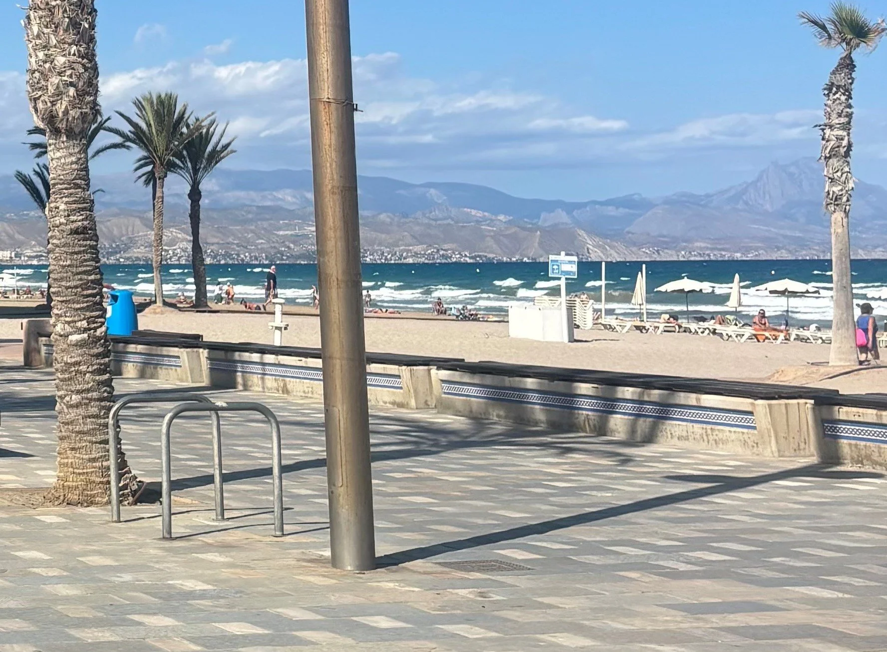 Beach scene with palm trees, ocean waves, beaches chairs, umbrellas, and mountains in the background.