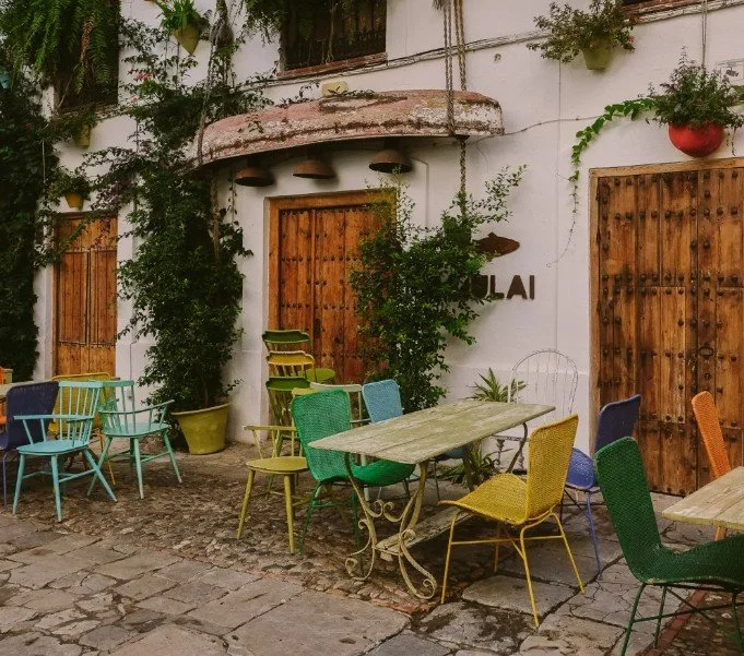 Outdoor seating area with colorful chairs and tables outside a building with wooden doors and greenery.