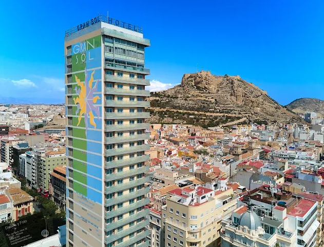 Tall hotel building with colorful signage that reads 'Grand Sol Hotel,' overlooking a cityscape with a hill in the background.